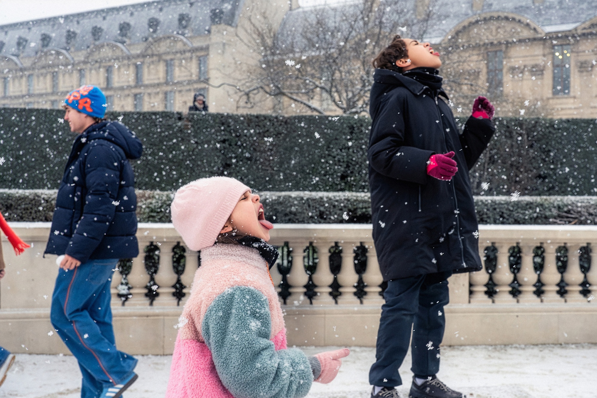 Children in coats and gloves stand outdoors catching snowflakes on their tongues as an adult walks by; snow falls, with a historic building visible in the background.