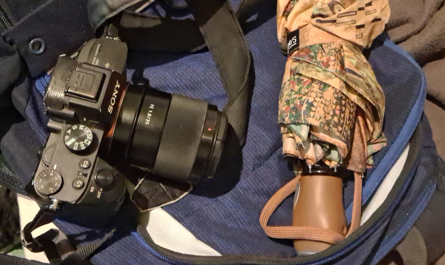 Sony camera with attached lens and a folded patterned umbrella placed on a blue and black bag.