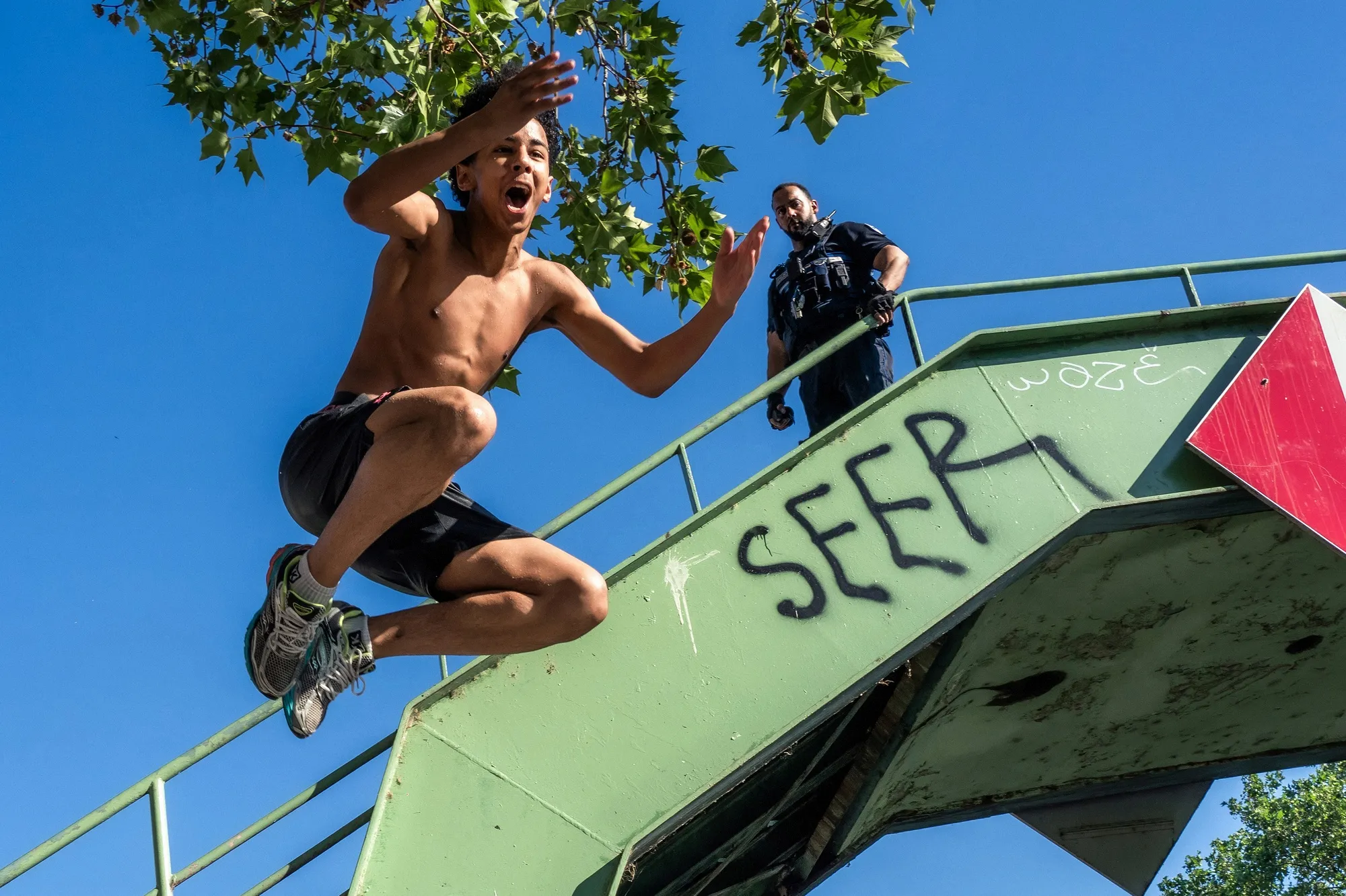 A person jumps off a green structure displaying SEER graffiti, while another individual observes from above; blue sky and green leaves in the background.
