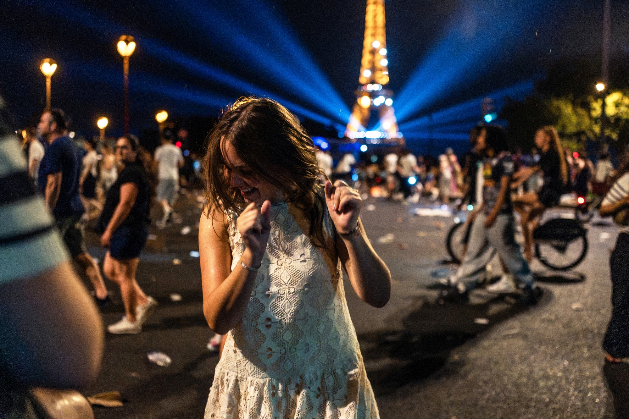 Person in a white dress dancing at night near the Eiffel Tower, which is lit with blue lights; people and a cyclist are in the background, and litter is scattered on the street.