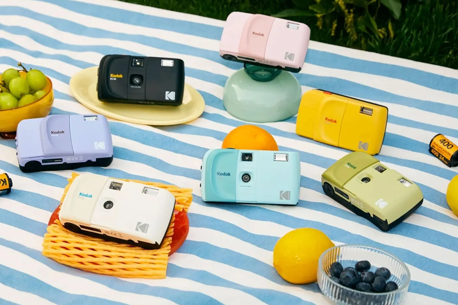 Six Kodak cameras in various bright colors rest on a blue-and-white striped blanket outdoors, with assorted fruit and several Kodak film rolls surrounding them.